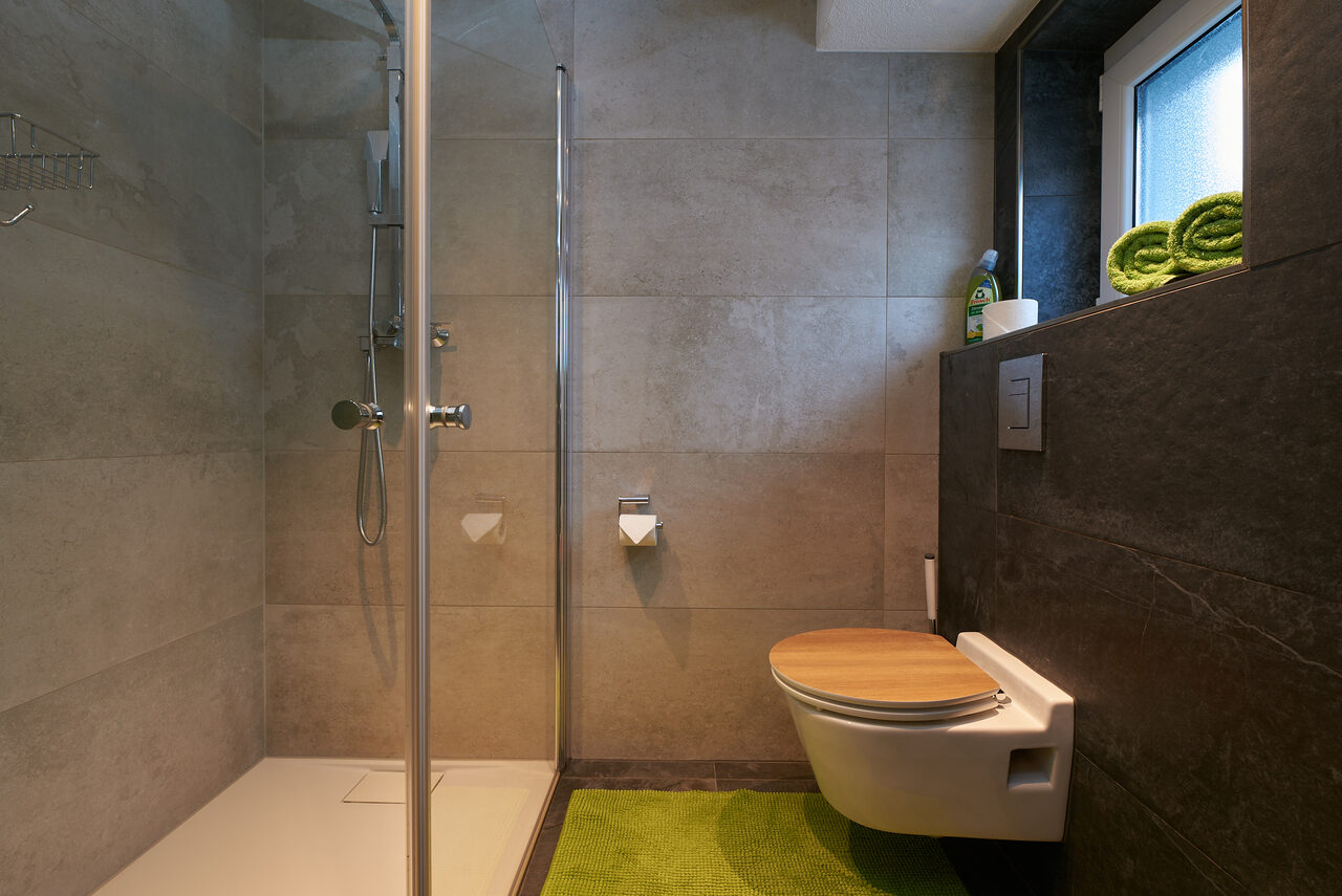 Bad mit bodengleicher Dusche hinter Glas, WC und einer Nische mit grünen Handtüchern.