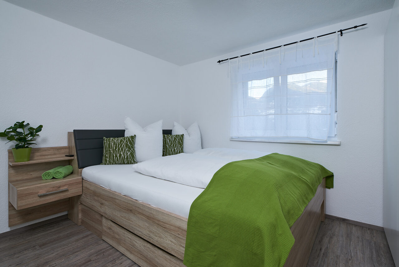 Doppelbett mit heller Bettwäsche und grüner Tagesdecke vor einer weißen Wand, daneben ein Fenster.